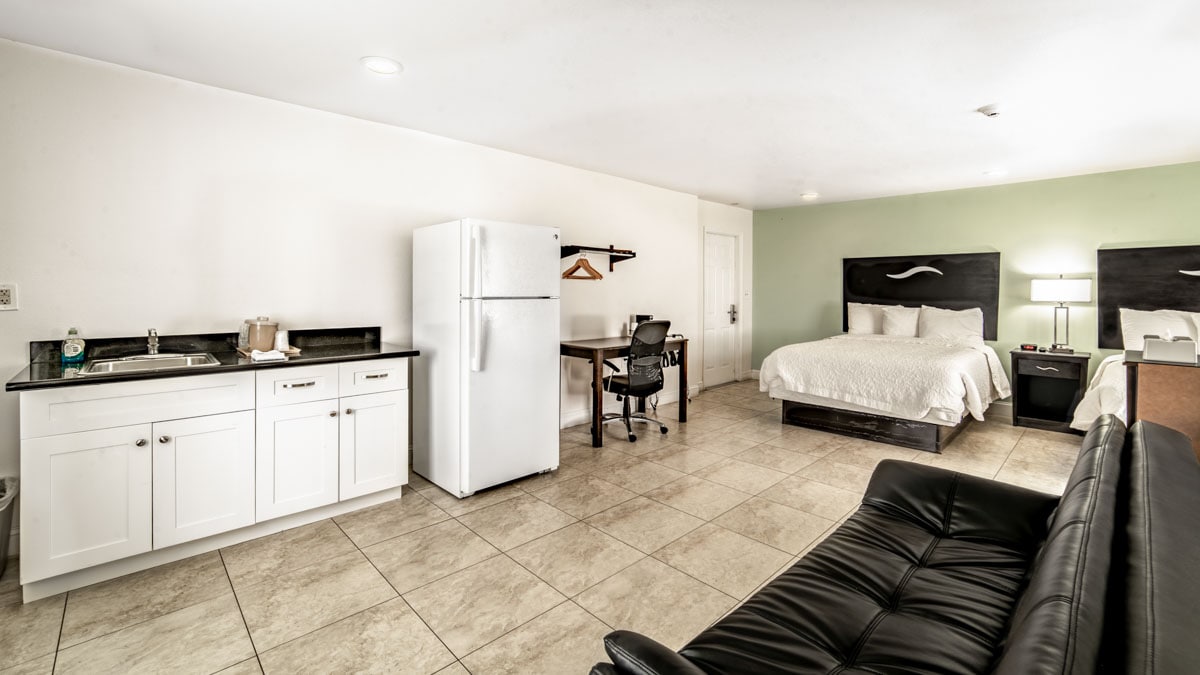 An efficiency room features two queen beds with soft bedding and a modern design. A kitchenette area is equipped with a full-size refrigerator and a sink. A work desk with a chair is positioned beside a sofa, creating a functional living space.