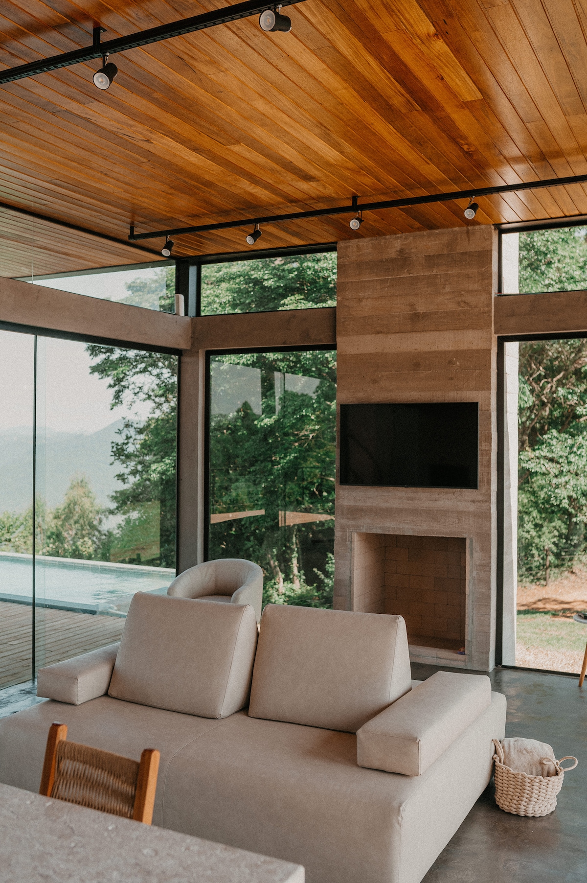 A modern living area showcases a light-colored sofa arranged near a fireplace and a wall-mounted television. Expansive windows invite natural light and provide views of the surrounding greenery. The wooden ceiling adds warmth to the space, creating a comfortable ambiance.