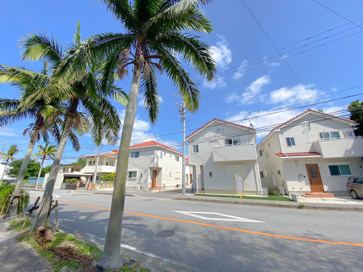 Two modern villas are positioned side by side along a palm-lined street. The white exteriors are complemented by red-tiled roofs, and clear blue skies provide a bright backdrop. A glimpse of a landscaped garden is visible in front of the properties.
