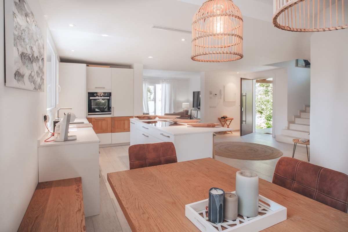 An open-plan kitchen and dining area blends functionality and style. A wooden dining table with leather chairs is positioned near modern pendant lights. The kitchen features sleek cabinetry and built-in appliances, while large windows allow ample natural light to fill the space, enhancing the airy feel.