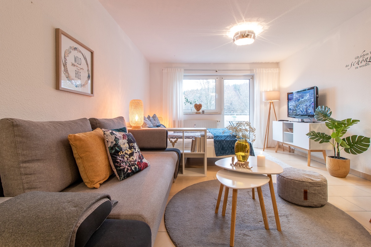 A bright living space features a comfortable grey sofa adorned with decorative pillows. A coffee table sits in front of the sofa, complemented by a modern rug. A flat-screen TV is mounted on a light-colored media unit, with natural light filtering in through a large window.