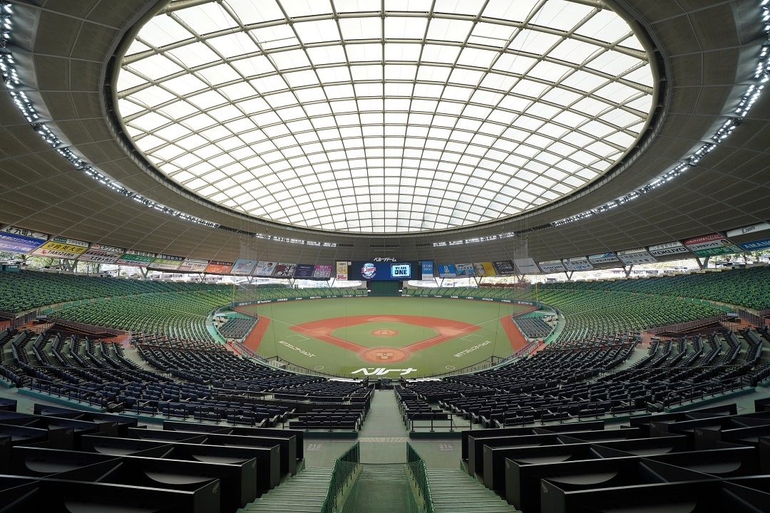 An expansive view of a stadium is captured from a high vantage point, showcasing the well-maintained green field surrounded by rows of seating. The distinctive dome-shaped ceiling allows natural light to illuminate the interior, creating an open and airy atmosphere.