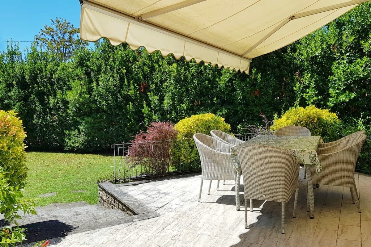 A shaded outdoor terrace features a round dining table surrounded by wicker chairs. Lush green hedges frame the area, providing privacy. The paved surface leads to a well-maintained lawn, with colorful shrubs adding a touch of vibrancy to the setting.
