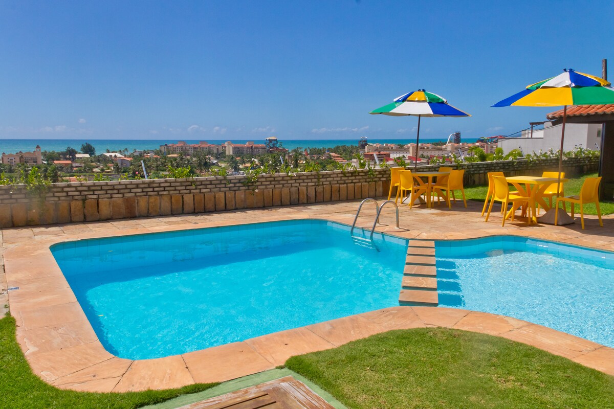 A private pool is surrounded by sun loungers and colorful umbrellas, offering a relaxing outdoor space. The view includes the ocean and nearby buildings, with clear blue skies overhead. A dining area with yellow chairs is positioned beside the pool, enhancing the serene environment.