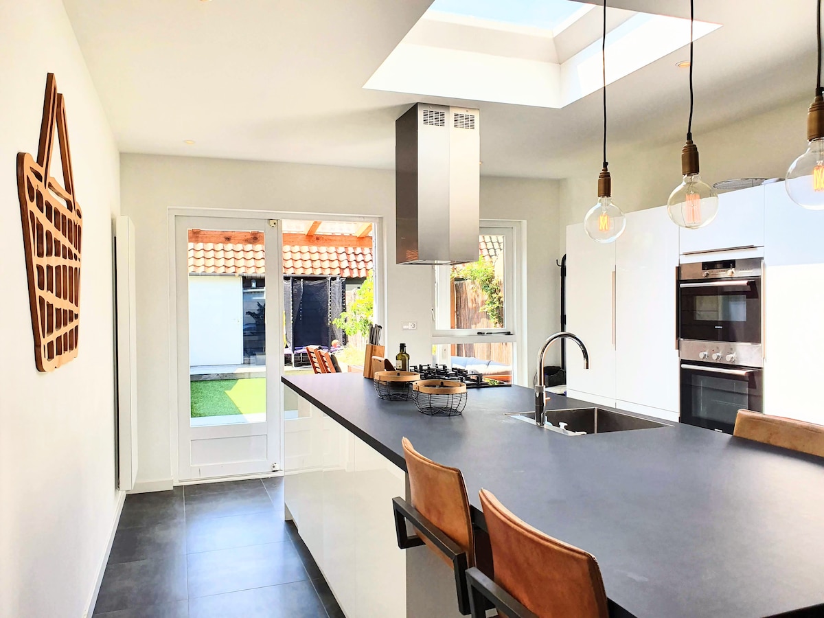 An open kitchen features a dark countertop and modern appliances. Natural light enters through a skylight and large glass doors, which lead to a garden area. The space includes bar seating with wooden stools and a decorative wall basket enhances the design.