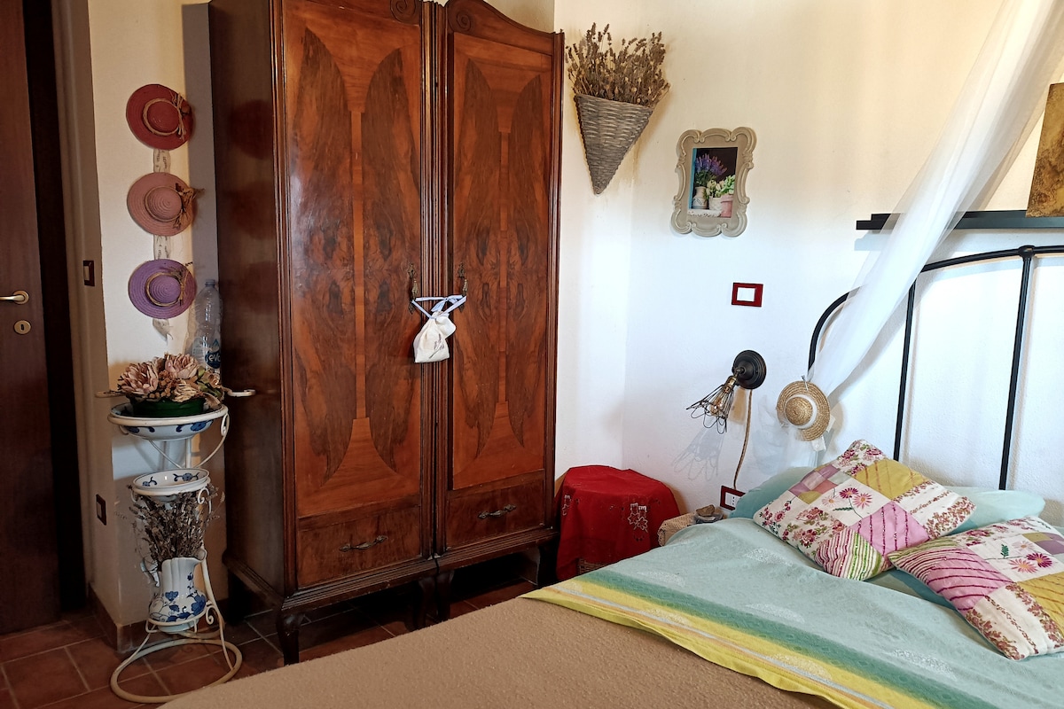 A cozy bedroom features a spacious bed with a light-colored blanket and patterned cushions. Beside it, a vintage wardrobe provides storage. Wall decorations include hats and a floral arrangement. A small table with a decorative lamp adds to the intimate atmosphere.
