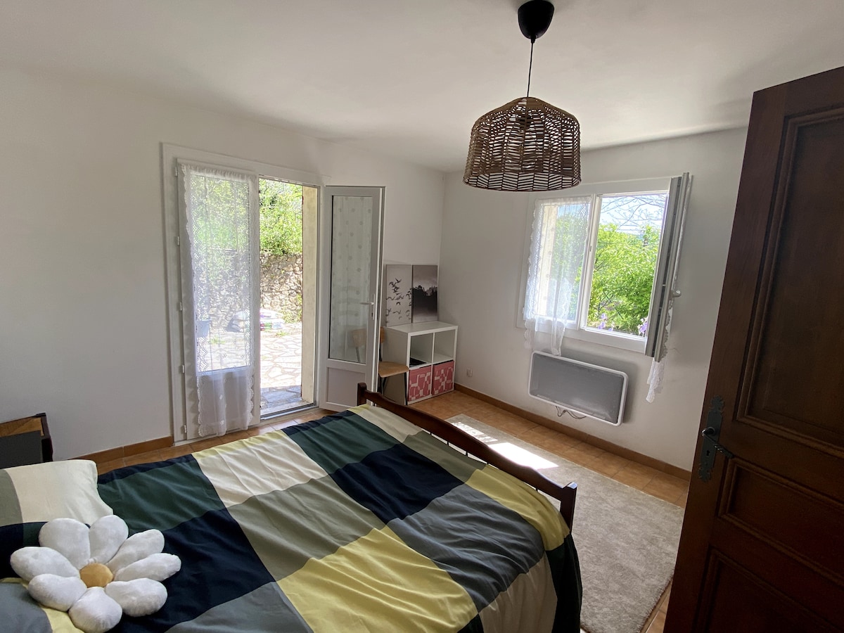 A bedroom features a spacious bed adorned with a patterned blanket, complemented by plush pillows. Natural light enters through a window and glass door, which leads to an outdoor area. A decorative basket pendant light hangs above, creating a bright and airy atmosphere.
