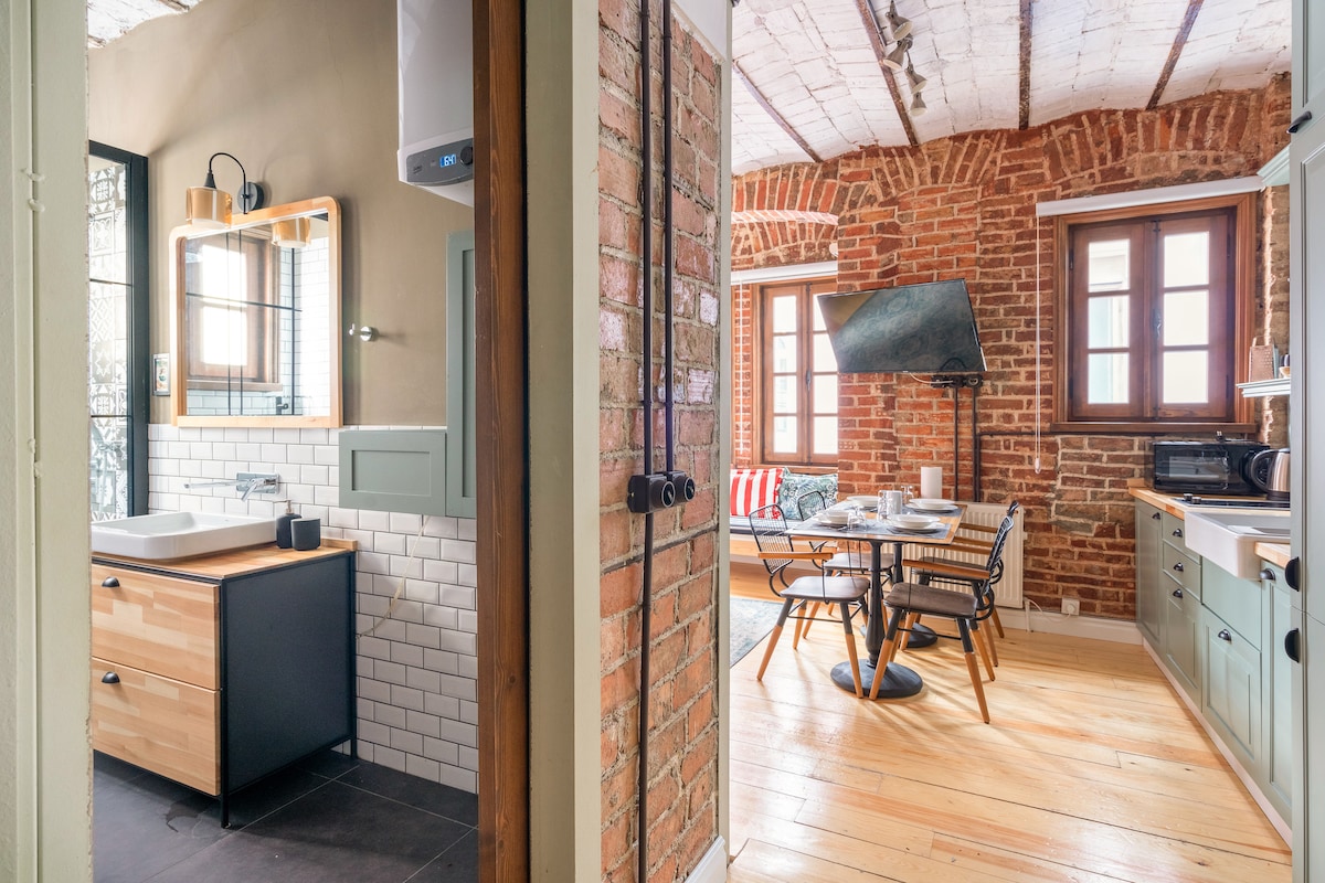 The image captures the entrance to the apartment, showcasing a blend of modern and historical design. A rustic brick wall contrasts with sleek furniture in the kitchen area, featuring a dining table set for four. The bathroom is visible with minimalist cabinetry and a bright mirror.