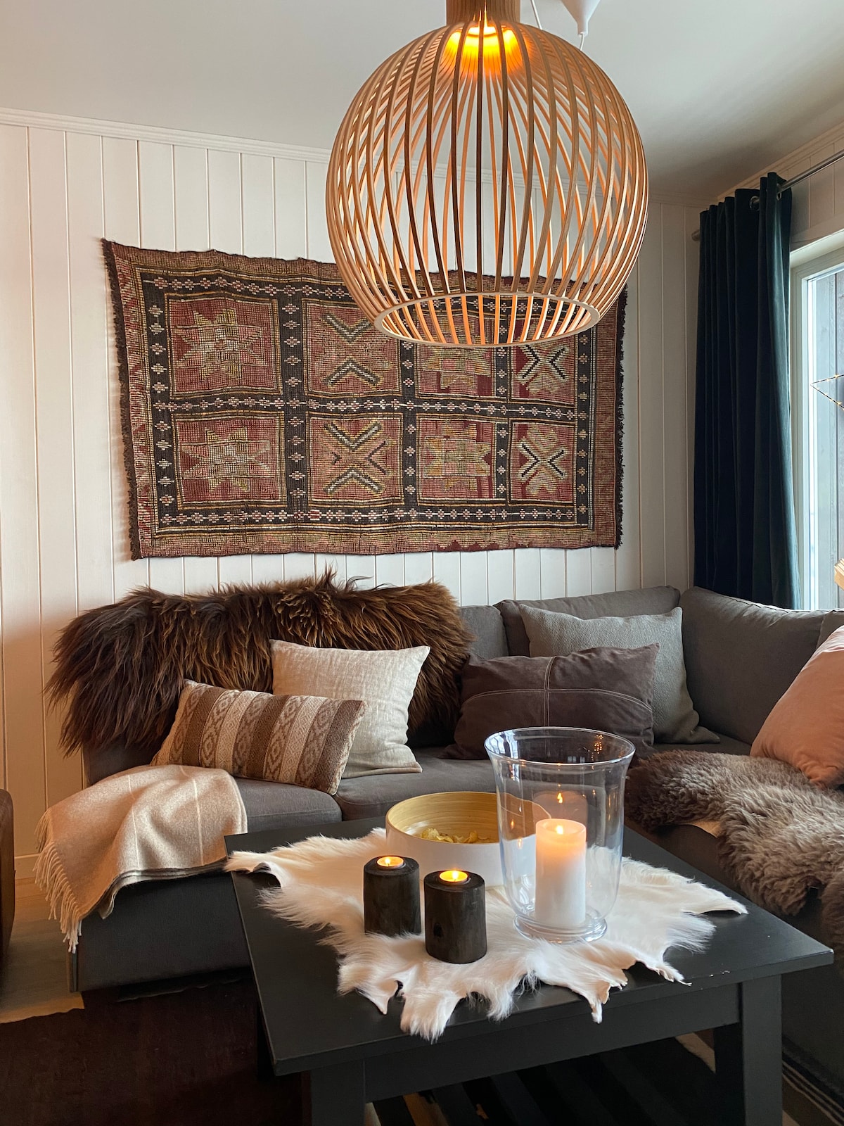 A cozy living area features a spacious gray sofa adorned with various textured cushions and soft throws. A woven tapestry hangs on the wall, complemented by a coffee table that holds candles and a clear vase. Natural light streams through a window dressed with dark curtains.