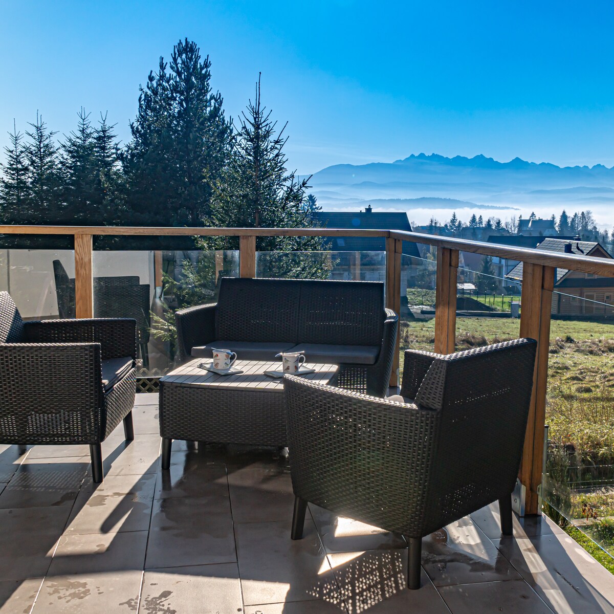 A balcony area features four comfortable, dark wicker chairs arranged around a glass-top table. The space is surrounded by trees, with expansive views of the mountains and sky in the background, reflecting a serene outdoor environment.