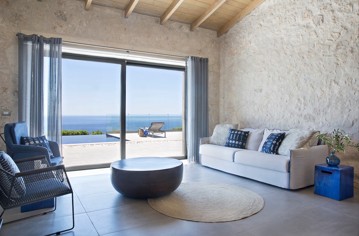 A spacious living area features large sliding glass doors that open to a panoramic view of the sea. Natural light floods the room, highlighting stone walls and wooden beams. A round coffee table is centered among a sofa and two accent chairs, creating a comfortable gathering space.