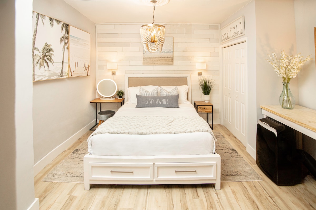 A serene sleeping area features a stylish bed with a light-colored headboard and decorative pillows. Soft lighting is provided by bedside sconces, while a modern chandelier hangs from the ceiling. A small desk and chair are positioned nearby, enhancing the functional space.