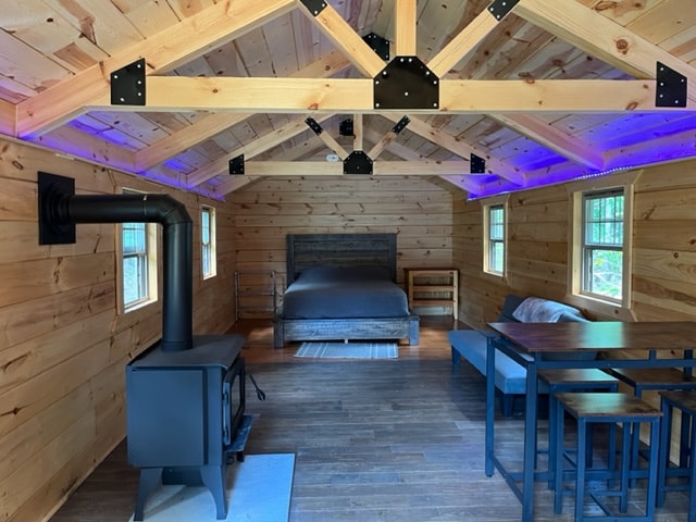 An interior view of the cabin features natural wood walls and a vaulted ceiling. A spacious Queen size bed is positioned against the back wall. A small wood stove is seen in the corner, while a dining area is set up with a table and chairs next to a comfortable seating area.