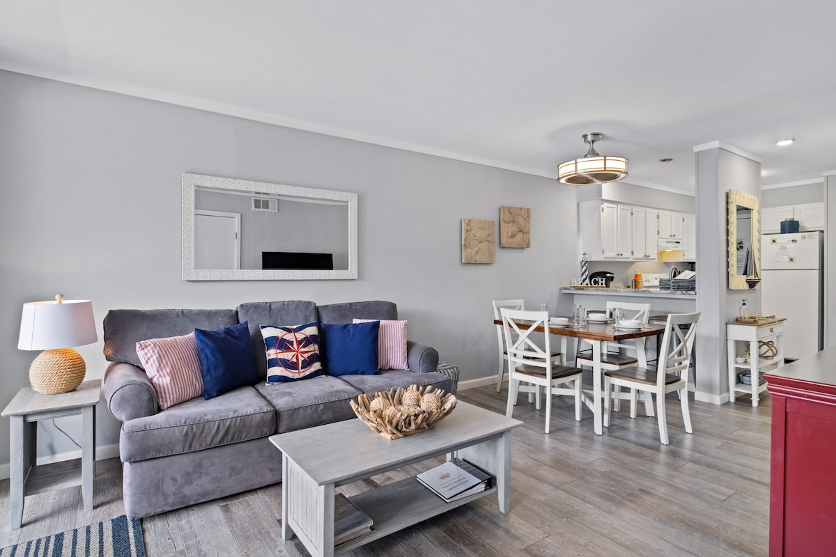 A comfortable living area features a gray sofa accented with colorful cushions. A wooden coffee table sits at the center, with a decorative bowl atop it. Adjacent, a dining table for four is visible, with a modern light fixture hanging above.
