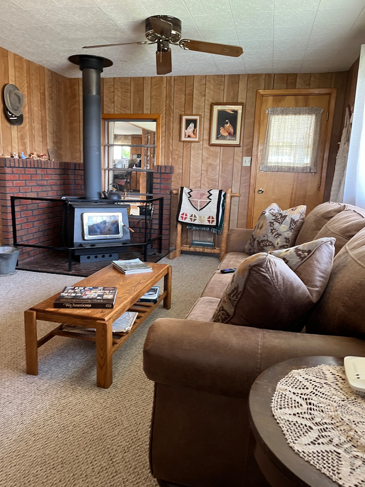 A cozy living area includes a plush sofa with decorative pillows and a wooden coffee table featuring magazines. A wood stove adds warmth, situated beside a rustic brick wall. Wall art and a handcrafted blanket draped over a chair enhance the inviting ambiance.