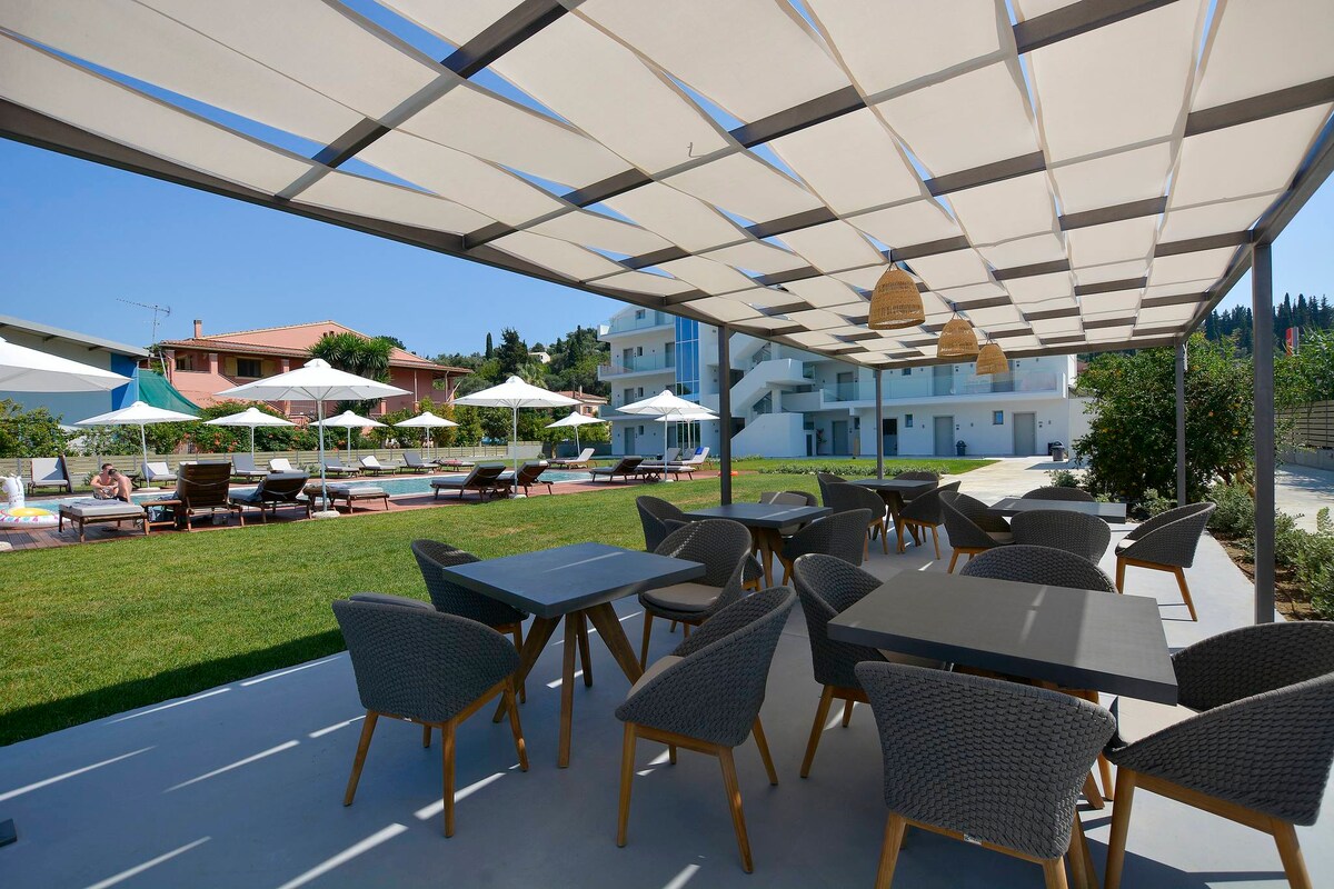 An outdoor dining area features modern wicker chairs surrounding rectangular tables. A shaded structure provides protection from the sun, while decorative pendant lights hang above. Lush green grass and a pool area are visible in the background, contributing to a relaxed ambiance.