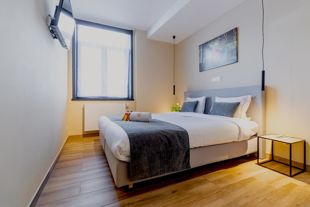 A well-lit bedroom features a spacious bed with neutral bedding and a soft throw. A small table beside the bed holds a towel and a decorative item. A television is mounted on the wall, and a framed artwork is displayed above the bed, offering a serene touch.