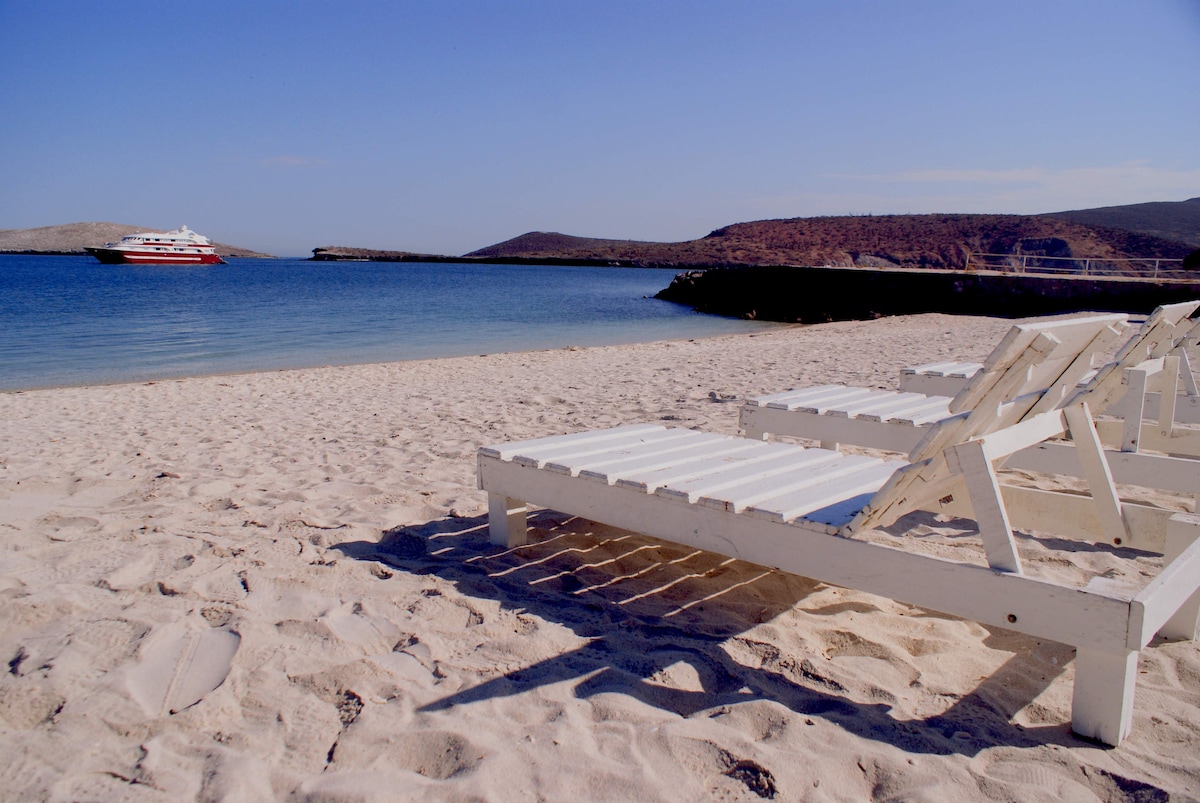 A serene private beach is visible, with white lounge chairs arranged on the soft sand facing the calm blue water. A boat is anchored in the distance, and gently rolling hills border the shoreline under a clear blue sky.