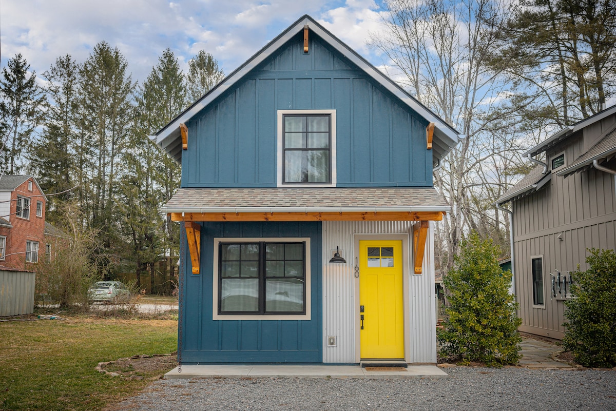 A charming two-story home features a blue exterior with a contrasting bright yellow front door. Large windows are framed in black, providing natural light to the interior. A gravel pathway leads to the entrance, surrounded by lush greenery and a grassy area.