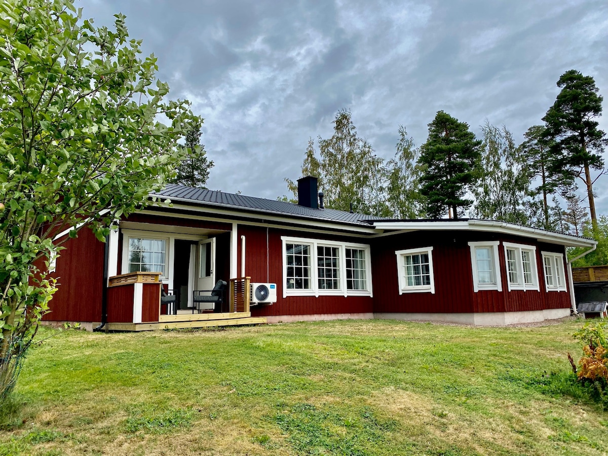 The spacious red villa is surrounded by greenery, featuring large windows that provide natural light. A wooden deck area with seating is visible, complemented by a chimney that adds a rustic charm. The setting reflects a peaceful landscape.