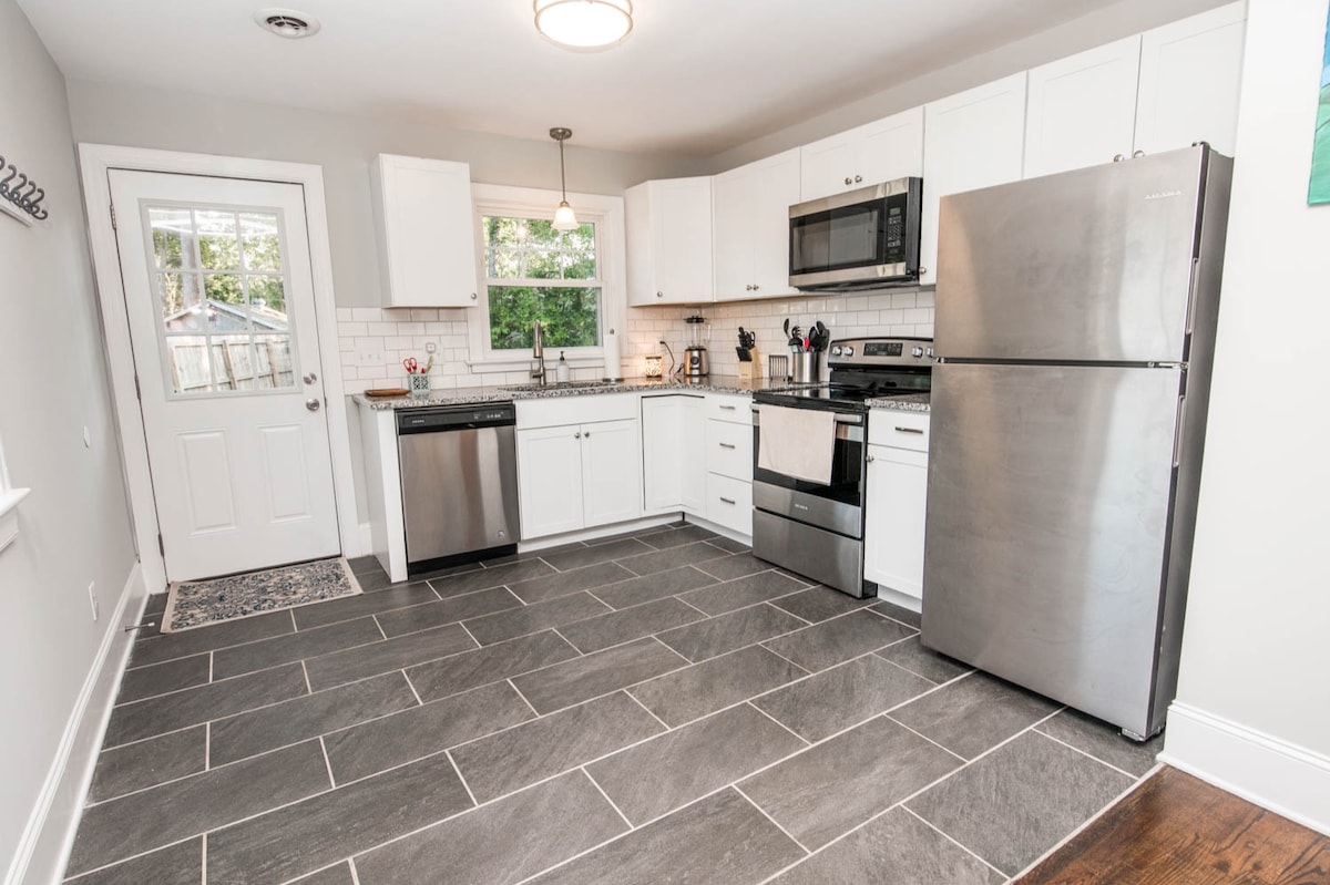A modern kitchen features stainless steel appliances, including a refrigerator, oven, and dishwasher. White cabinetry and a tiled backsplash create a clean aesthetic. Ample counter space is available for meal preparation, with a door leading to an outdoor area visible in the background.
