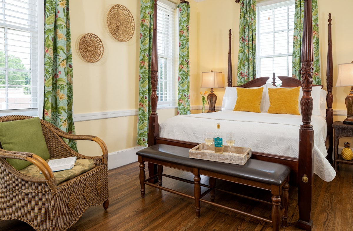 A bright and airy guest room features a four-poster bed with crisp white linens and yellow decorative pillows. Two bedside lamps provide soft lighting. A rattan chair with a cushion and a wooden bench with a tray sit in front of the bed, with patterned curtains framing the windows.
