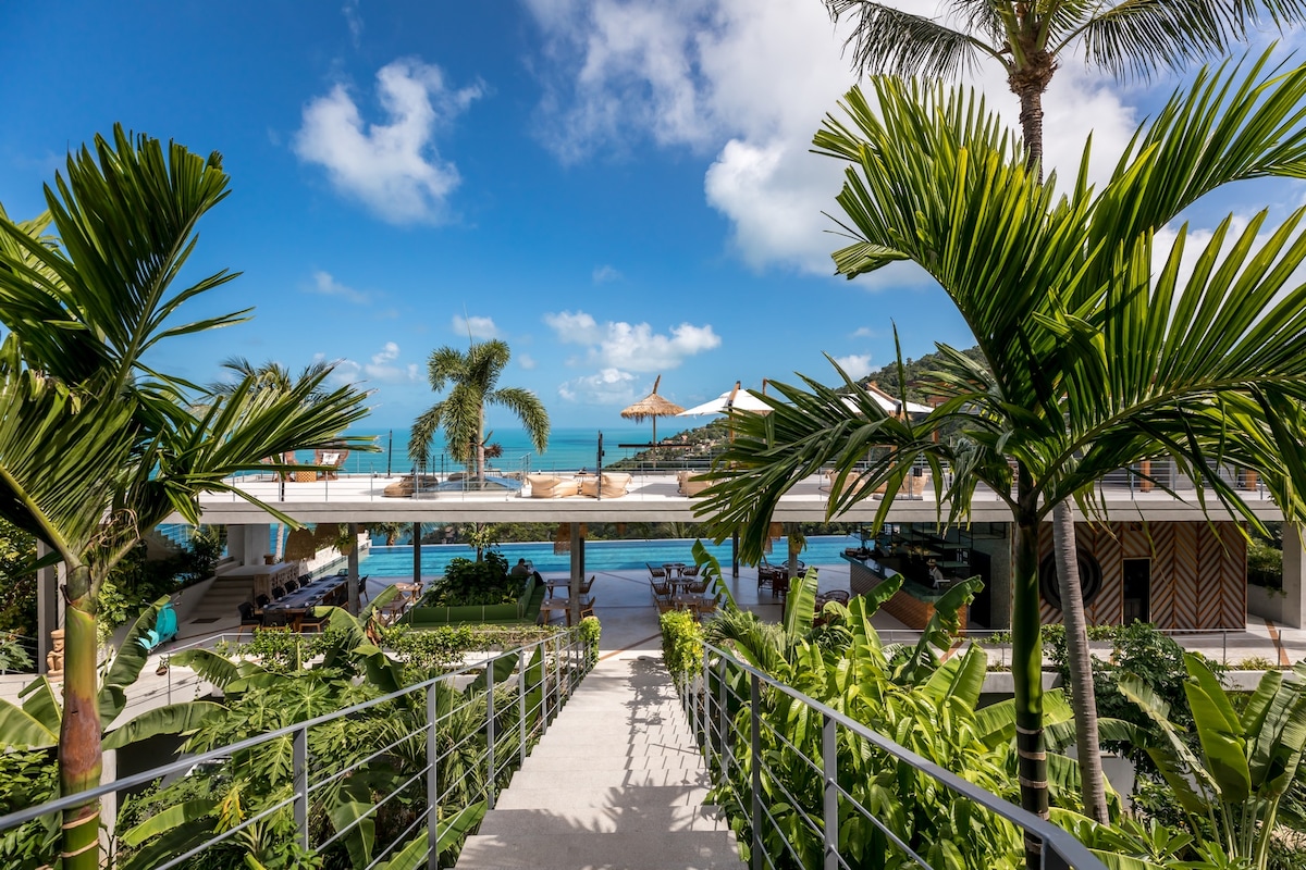 A pathway lined with tropical plants leads to a multi-level terrace area, featuring seating and sun umbrellas. The backdrop highlights a tranquil ocean view under a blue sky with scattered clouds, inviting relaxation and connection to nature.