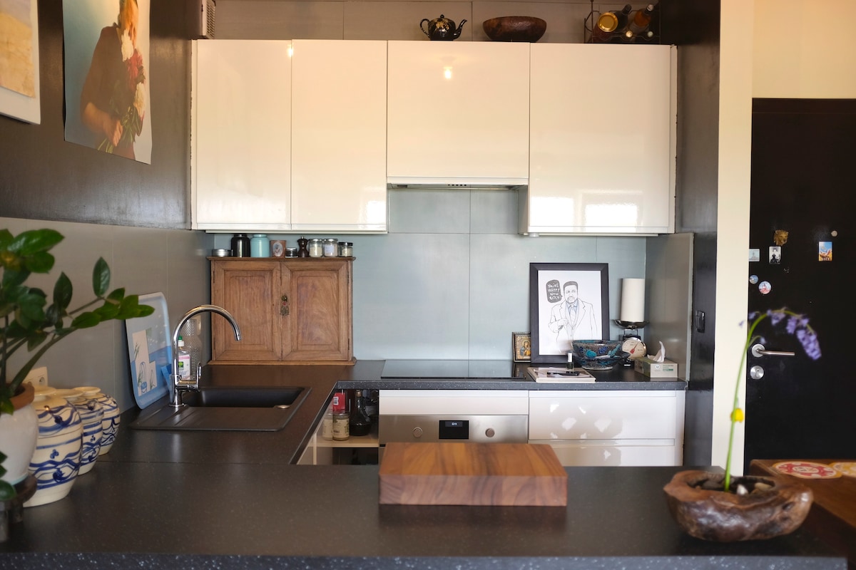A modern kitchen features sleek white cabinetry and a dark countertop. A wooden cutting board is placed prominently in front of the sink. Decorative items, including plants and art, are displayed on shelves, while a framed piece of art is set against the wall.
