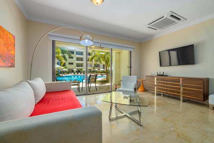 Salon relaxant avec vue sur la piscine. Cet appartement d'Aruba dispose d'un grand salon avec de grandes fenêtres offrant une vue sur la piscine centrale. L'espace aéré et élégant est parfait pour se détendre et profiter du paysage serein au bord de la piscine.