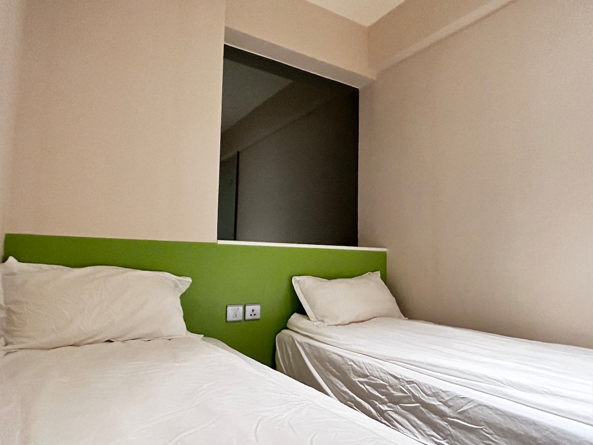 The room features two single beds positioned against a light green accent wall. The bedding is neatly arranged, and natural light enters through a window, casting soft shadows. The overall decor is simple and functional, providing a comfortable sleeping space.
