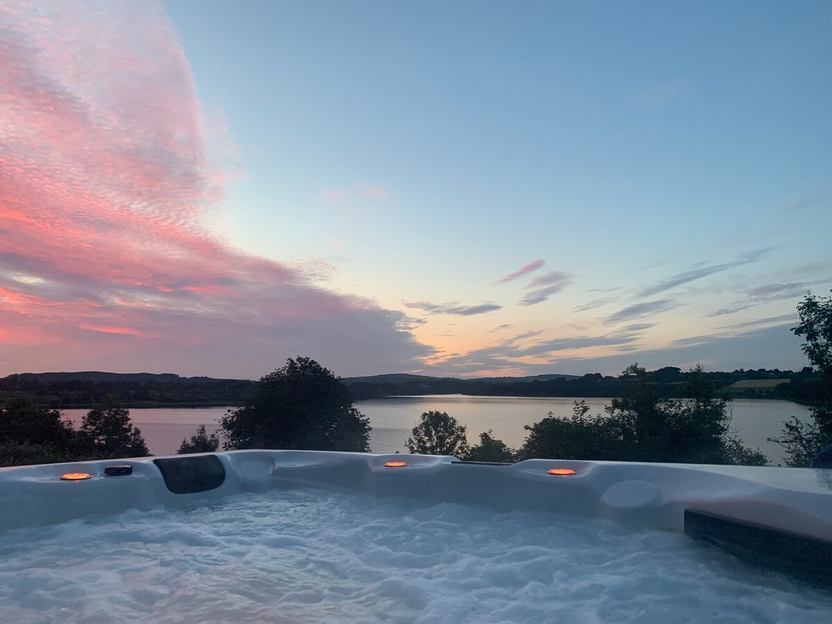 The hot tub is situated outdoors, with gentle steam rising from the water. A stunning sunset paints the sky in soft pinks and purples, creating a serene backdrop over Blessington lake. Lush greenery surrounds the area, enhancing the peaceful atmosphere.
