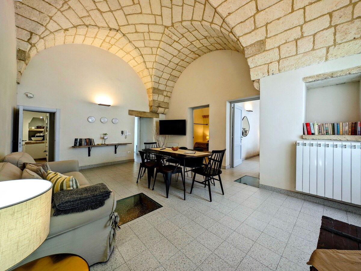 The spacious living area is characterized by high stone vaulted ceilings and modern furnishings. A comfortable sofa and dining table are set, with natural light illuminating the room through multiple open doorways. A section of the floor features glass panels, adding a unique design element.