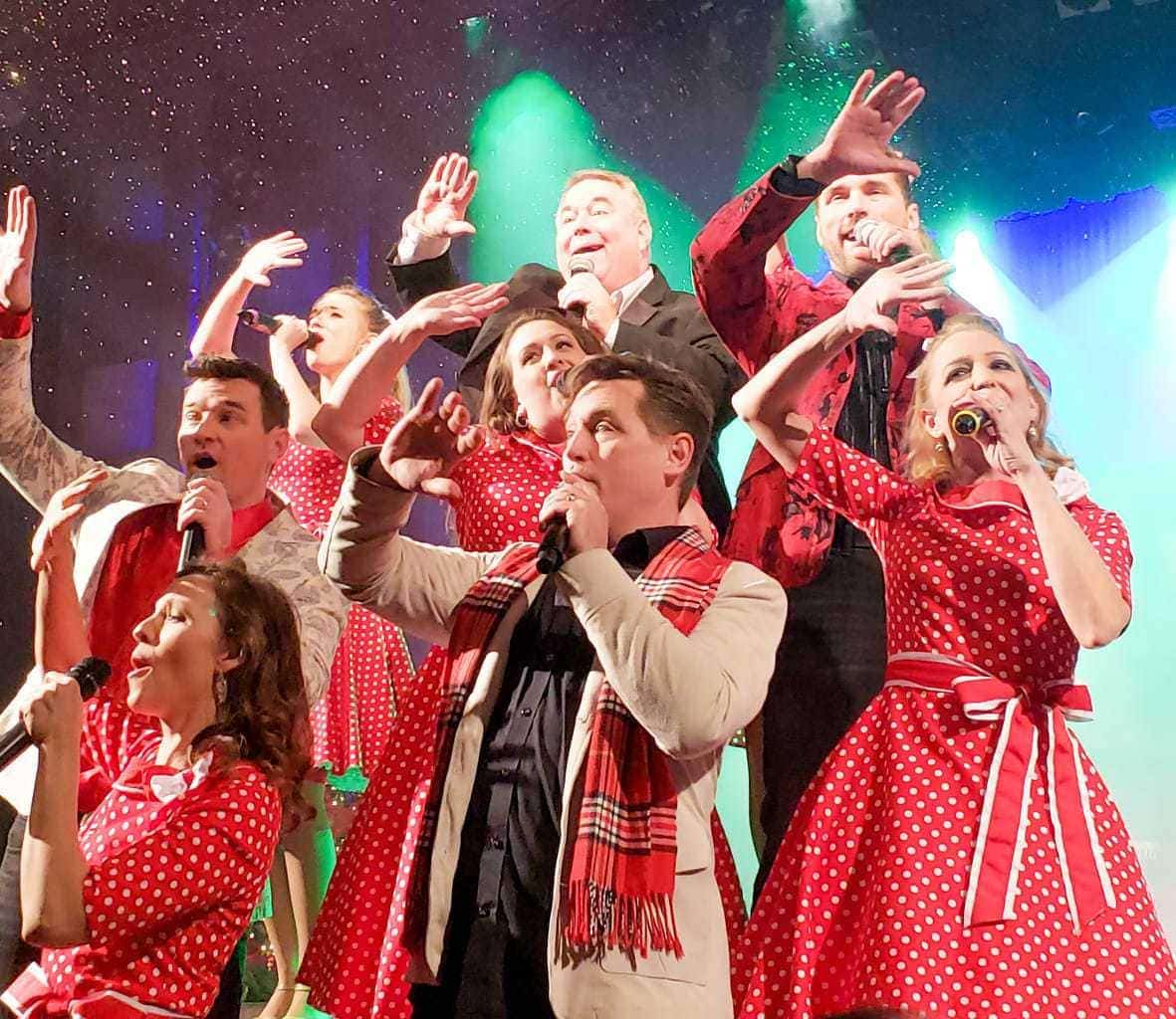 A vibrant performance stage features a group of entertainers dressed in festive attire, with polka-dot dresses and coordinated accessories. Various poses are struck against a colorful backdrop, highlighting a joyful atmosphere enhanced by theatrical lighting and sparkling effects.