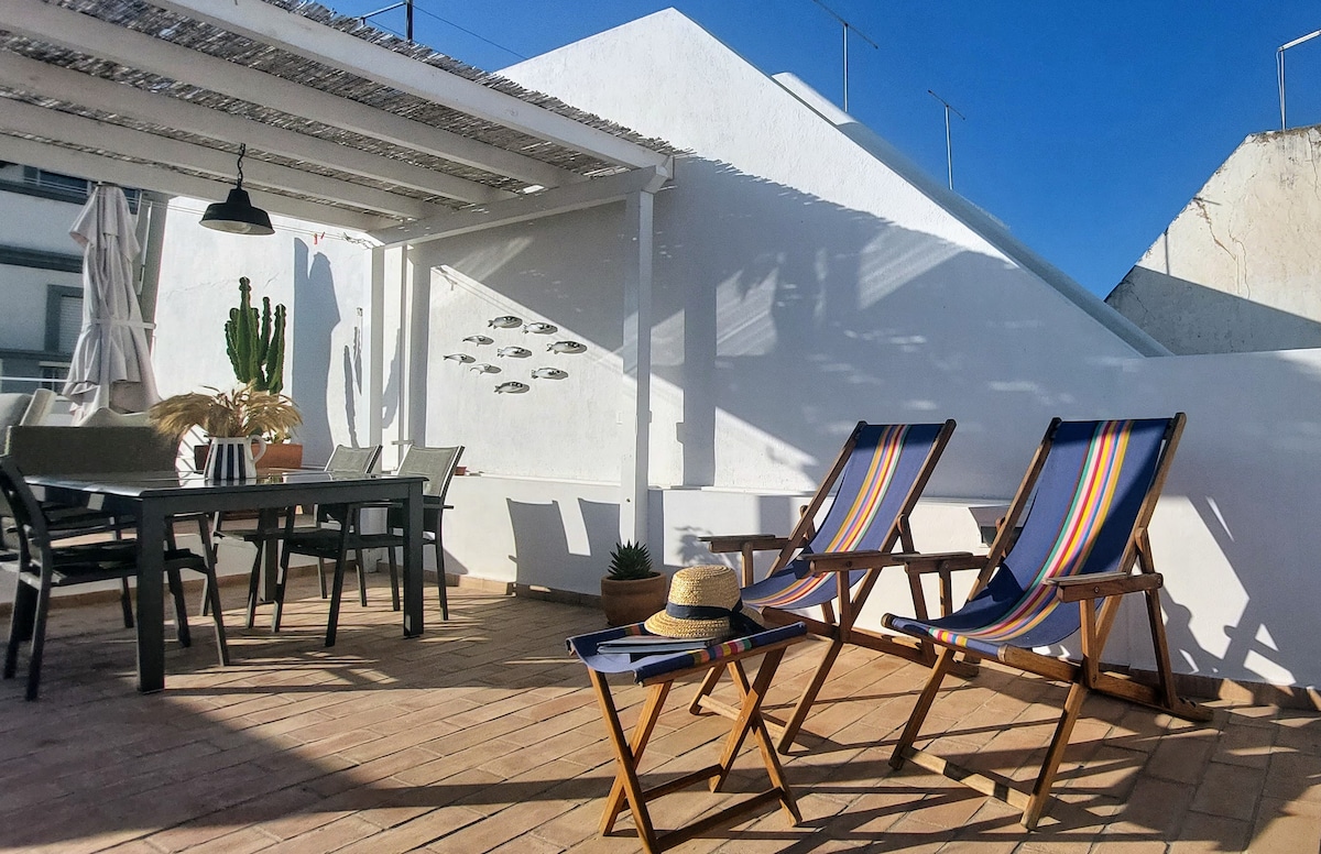 A rooftop terrace features a dining set along with two loungers positioned for relaxation. Sunlight illuminates the space, highlighting light-colored walls and a clear blue sky. Potted plants and decorative elements add a touch of greenery to the setting.