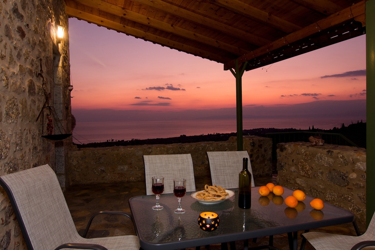 An outdoor terrace is illuminated by a soft glow, featuring a dining table set with wine glasses, a bottle, and a platter of snacks. The scenic view displays a colorful sunset over the sea, with orange and purple hues in the sky.