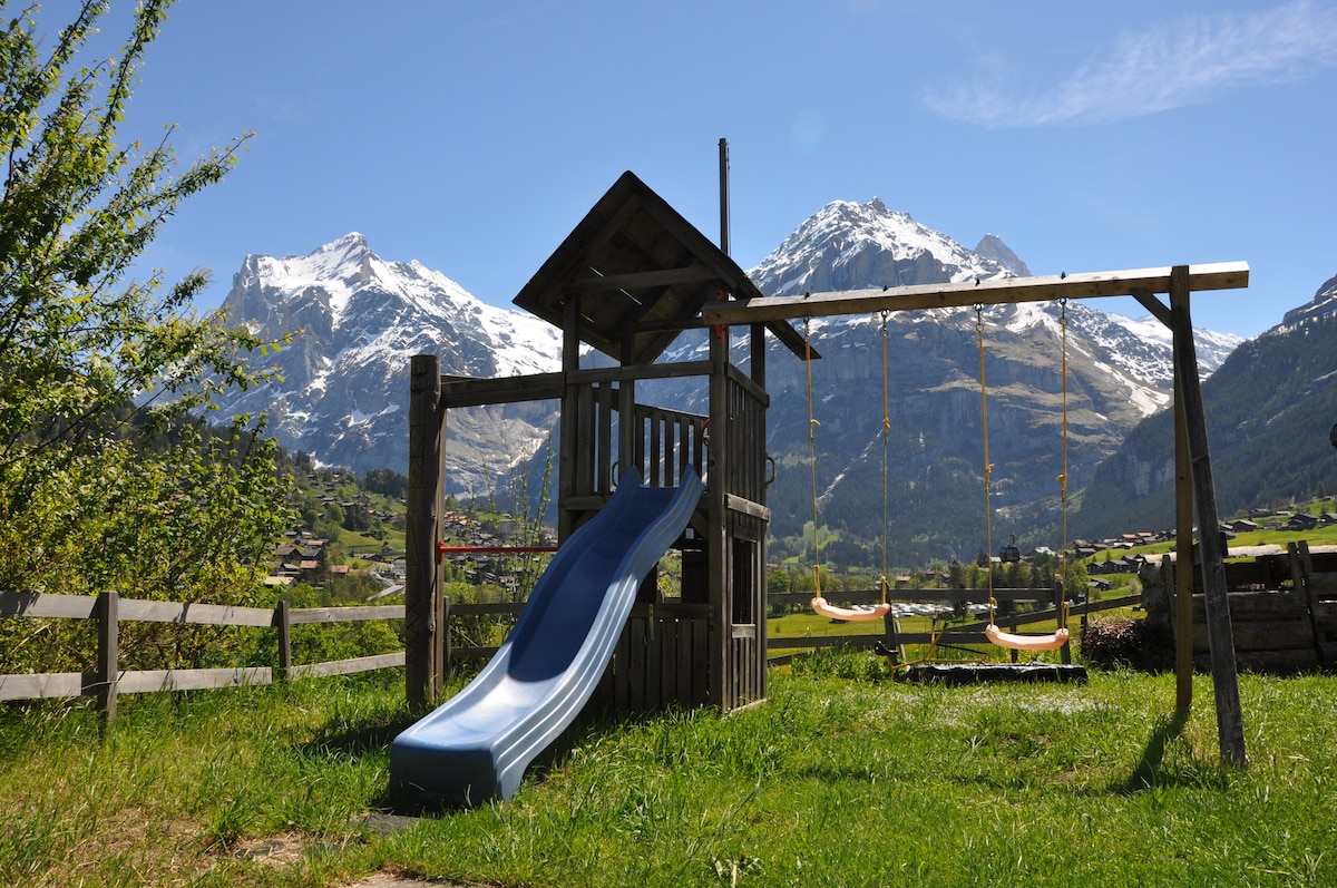 A wooden play structure features a slide and swings, set against a backdrop of majestic mountains under a clear blue sky. Lush green grass surrounds the play area, inviting outdoor activities.
