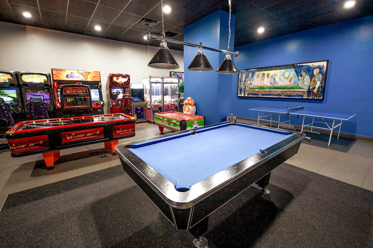 A game room is presented featuring various arcade machines along one wall, including a vibrant air hockey table. A blue felt pool table occupies the center space, illuminated by three overhead pendant lights. The walls are painted in a rich blue hue, enhancing the lively atmosphere.