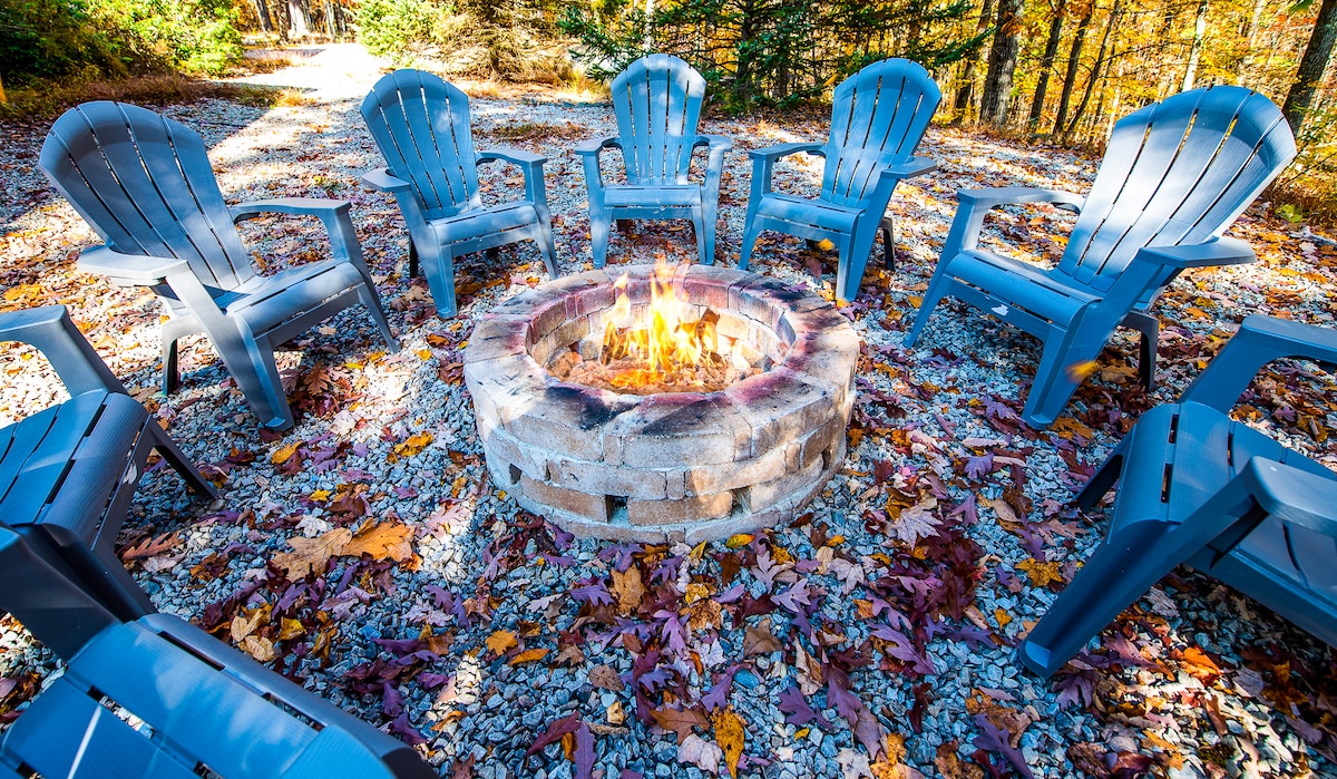 A circular fire pit area is surrounded by eight blue adirondack chairs, positioned on a gravel setting. Autumn leaves scatter across the ground, showcasing a blend of vibrant colors among the trees in the background.