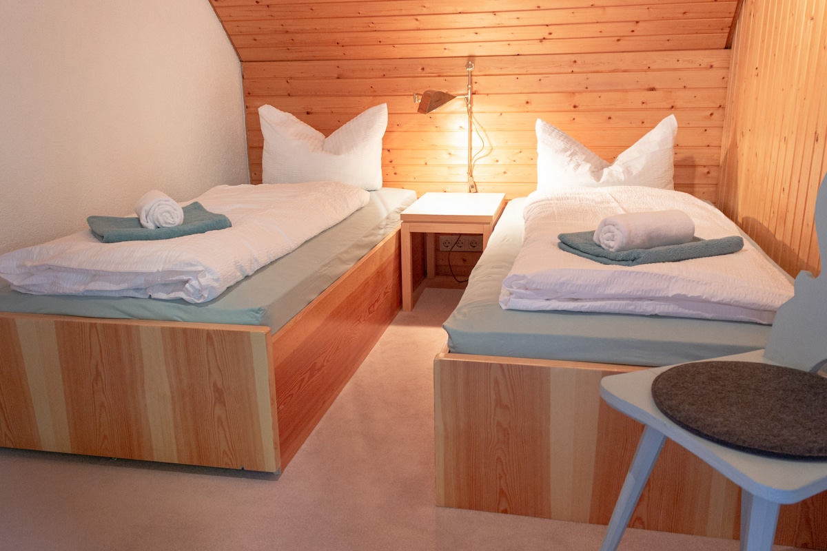 The cozy bedroom features two spacious beds with crisp white linens and decorative pillows. A bedside table is positioned between the beds, and a modern lamp provides soft lighting. The wooden paneling on the walls adds a warm touch to the tranquil atmosphere.