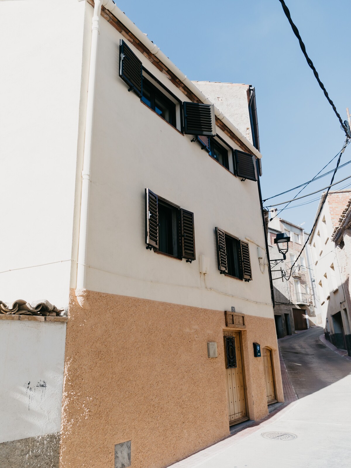 A three-story building with a textured beige and white façade is depicted. The front entrance is framed by wooden shutters on the upper windows. The street is narrow, featuring traditional architecture and overhead cables, with a clear blue sky above.