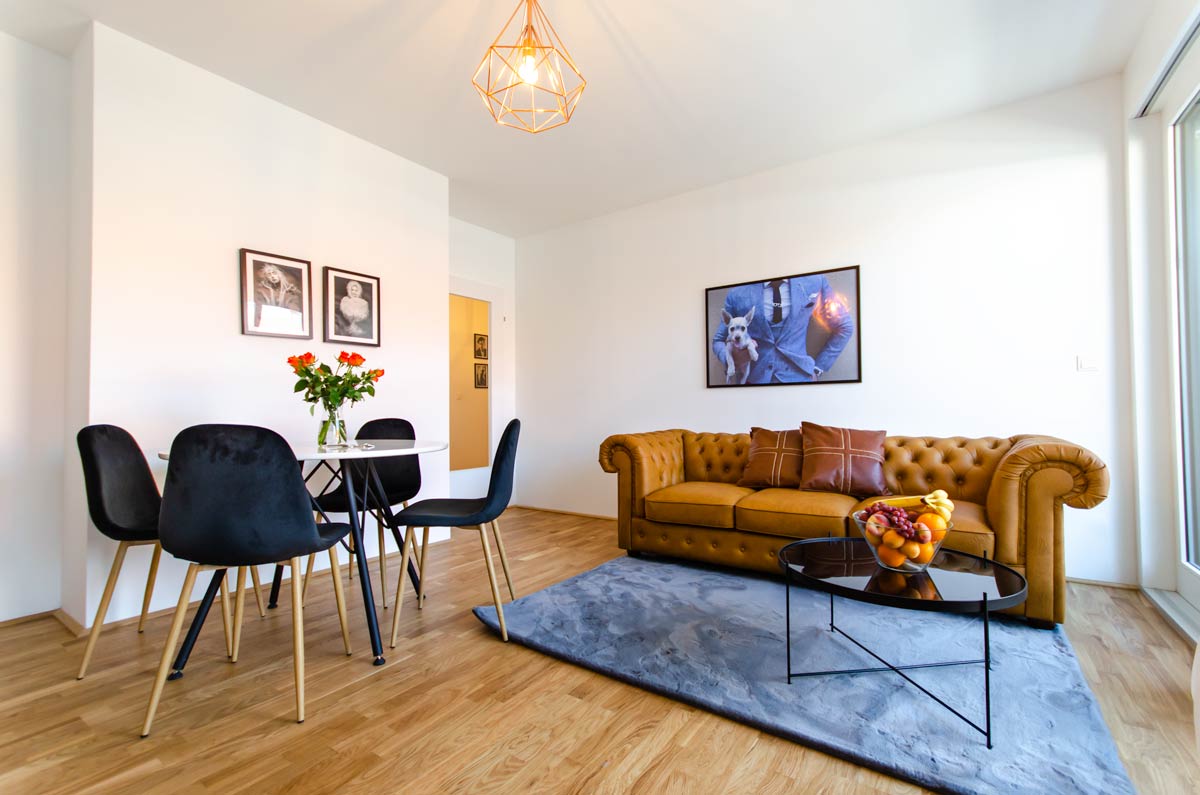 A bright living area features a leather sofa with decorative cushions and a round coffee table set above a soft area rug. A dining table with four stylish chairs is positioned nearby, complemented by framed art on the walls and a vase of fresh flowers.