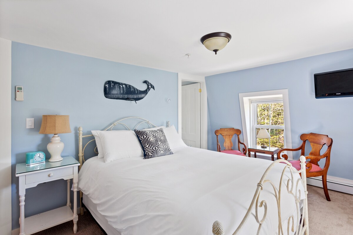 A bright bedroom features a spacious bed with a white quilt. Light blue walls are complemented by a decorative whale wall art. Two chairs are positioned near a window, which allows natural light to fill the room. A small bedside table includes a lamp and a turquoise trinket.