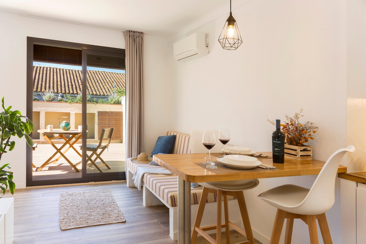 A light-filled dining area features a wooden table set for two, complemented by modern chairs. A cozy seating nook is styled with stripes and decorative pillows. Large glass doors open to a spacious terrace, creating a seamless connection with the outdoor space.