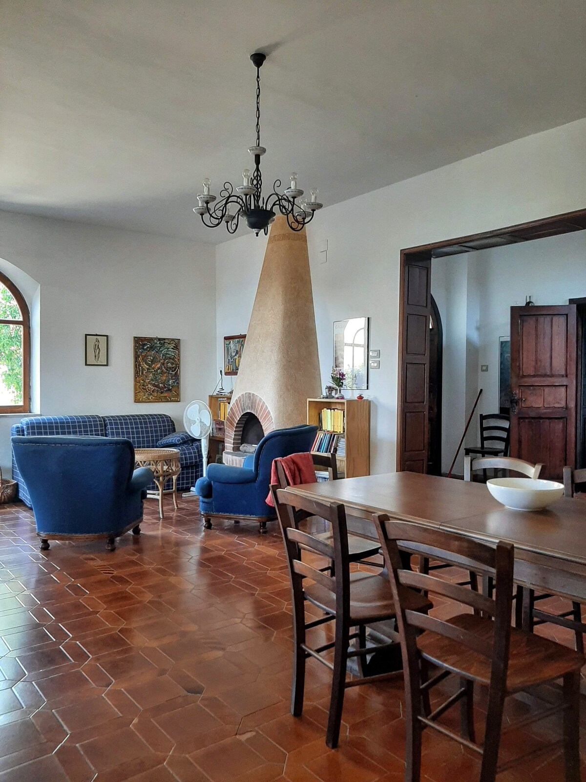 A spacious living area features a dining table surrounded by wooden chairs. An inviting fireplace stands at the center, complemented by a chandelier overhead. A sofa and armchairs provide seating options, enhancing the welcoming design. Natural light enters through arched windows, brightening the room.