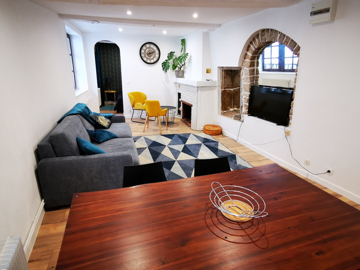 A living area features a gray sofa and a bright yellow armchair, arranged around a geometric-patterned rug. A wooden dining table sits in the foreground. A TV is mounted above a decorative fireplace, and stone accents are visible on the wall beside it.