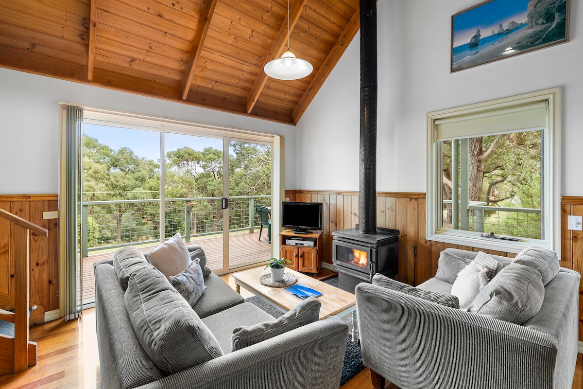 A warm living area features wood-paneled walls and a soaring ceiling with wooden beams. Two comfortable sofas are positioned around a coffee table, while a wood stove provides warmth. Large glass doors open to a deck with views of surrounding greenery.