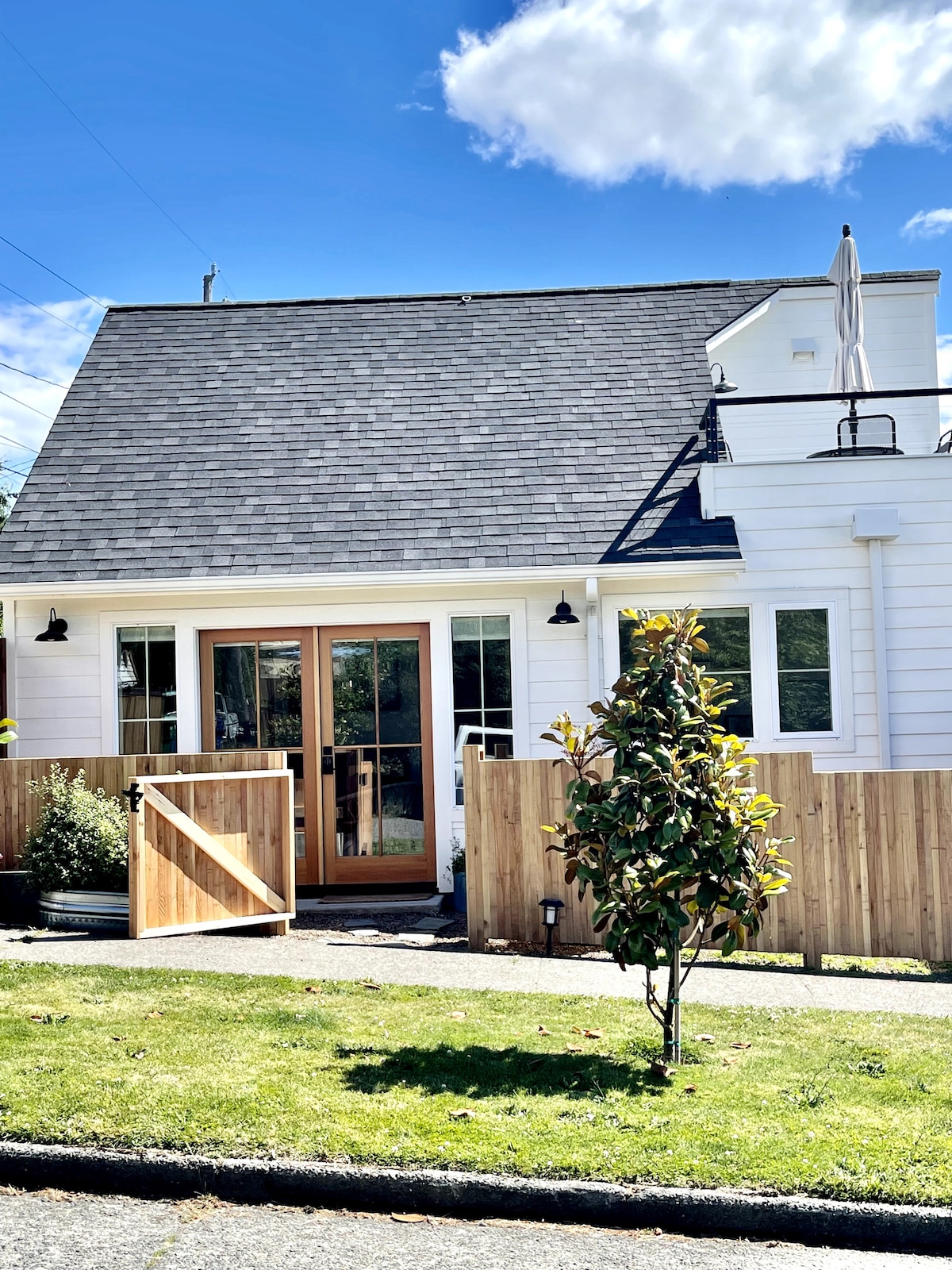 The exterior of the custom-built guesthouse features a sloped roof and welcoming wooden double doors. A small garden area is visible, complemented by a young tree and wooden fence. Blue skies and fluffy clouds provide a bright backdrop for this charming structure.
