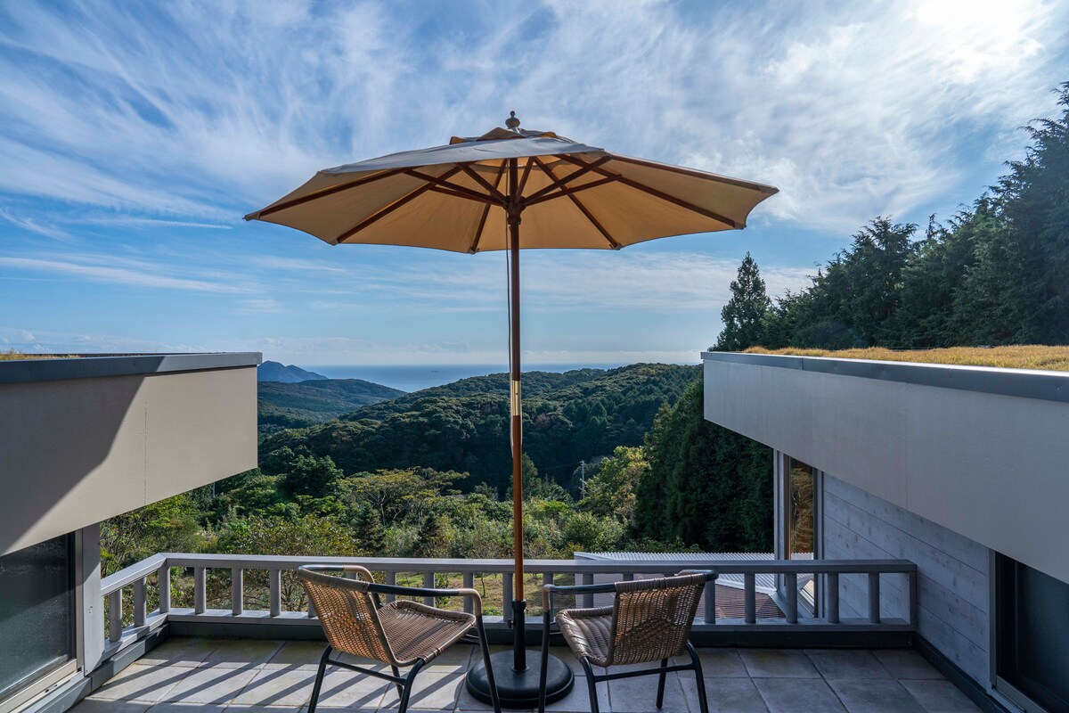 A shaded outdoor seating area features two wicker chairs positioned under a large umbrella. The view embraces rolling hills and a distant sea under a bright sky, creating a serene backdrop for relaxation.