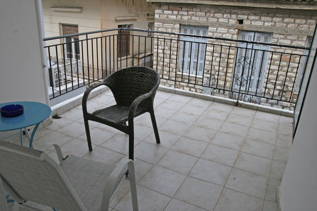 A small balcony features tiled flooring and a safety railing, overlooking a courtyard with stone walls. A wicker chair and a blue side table provide a cozy spot for relaxation. Sunlight reflects off the balcony, creating a bright and open atmosphere.
