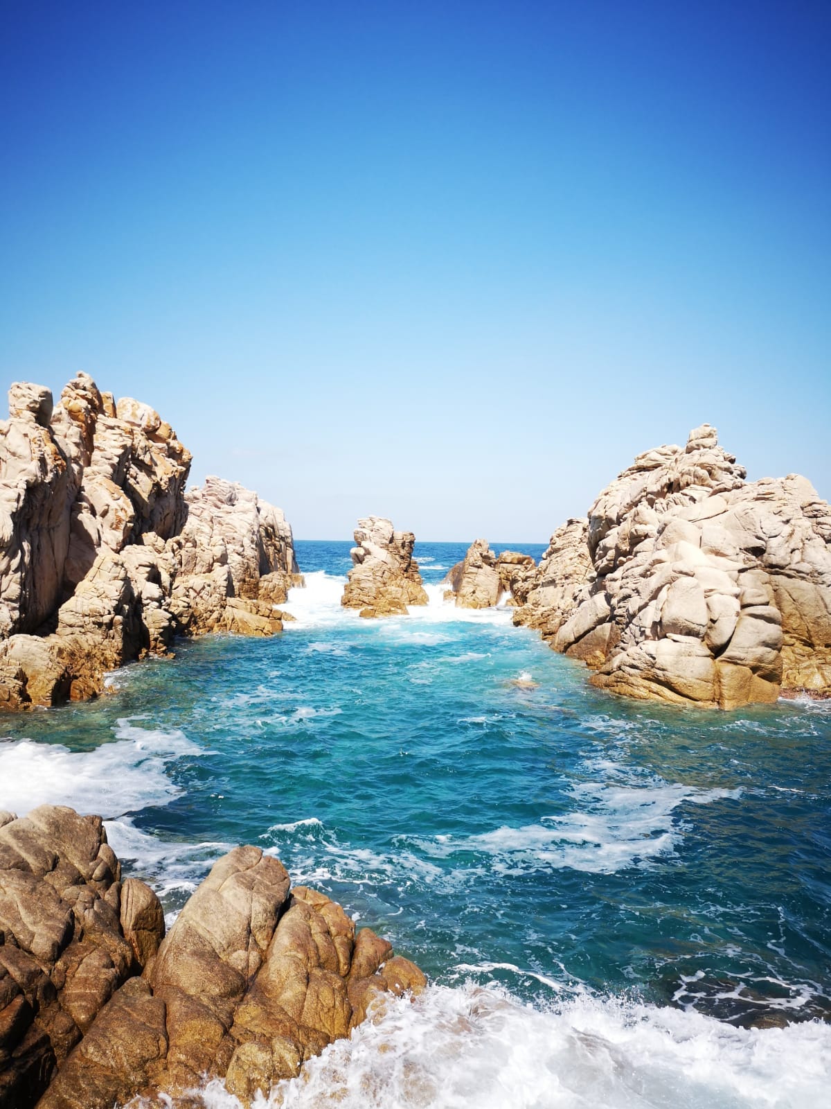 The image displays rocky cliffs emerging from clear blue waters, with waves gently lapping against the stones. A bright blue sky fills the background, creating a serene coastal scene, inviting exploration of the natural beauty surrounding the area.